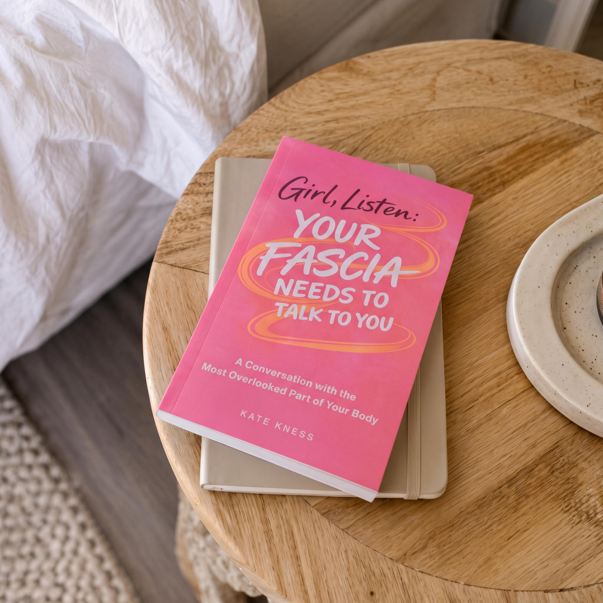 Pink paperback edition of Girl, Listen: Your Fascia Needs to Talk to You by Kate Kness, resting on a notebook on a round wooden table.